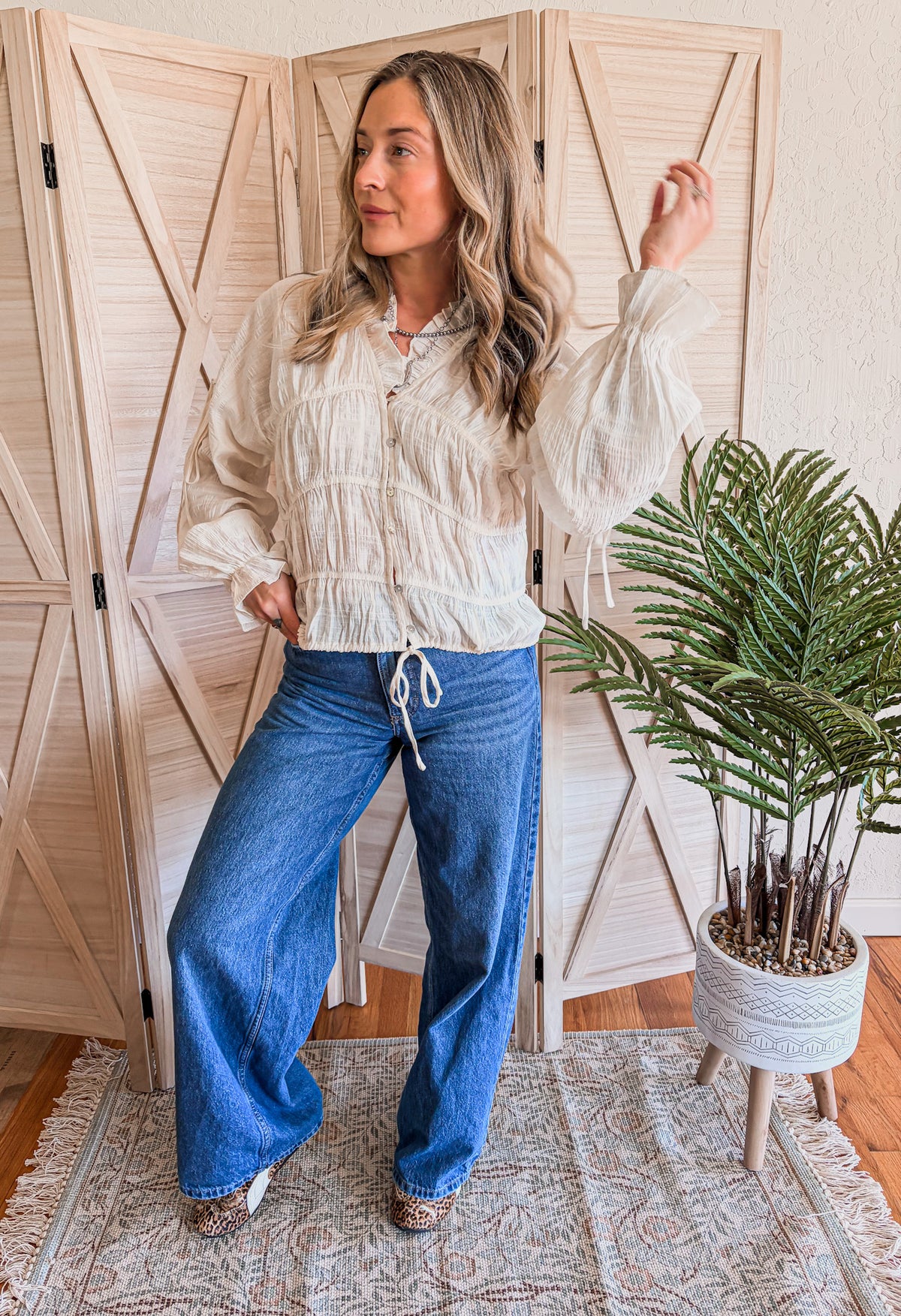 Woman wearing a cream blouse and blue jeans standing in a room with wooden shutters and a plant.