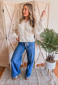 Woman wearing a cream blouse and blue jeans standing in a room with wooden shutters and a plant.