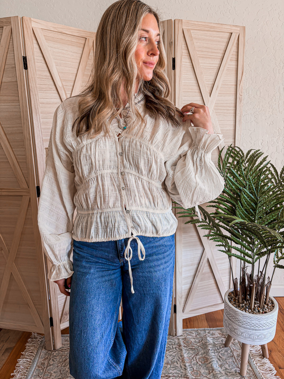 Woman wearing a cream blouse and blue jeans standing in a room with wooden shutters and a plant.