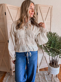 Woman wearing a cream blouse and blue jeans standing in a room with wooden shutters and a plant.