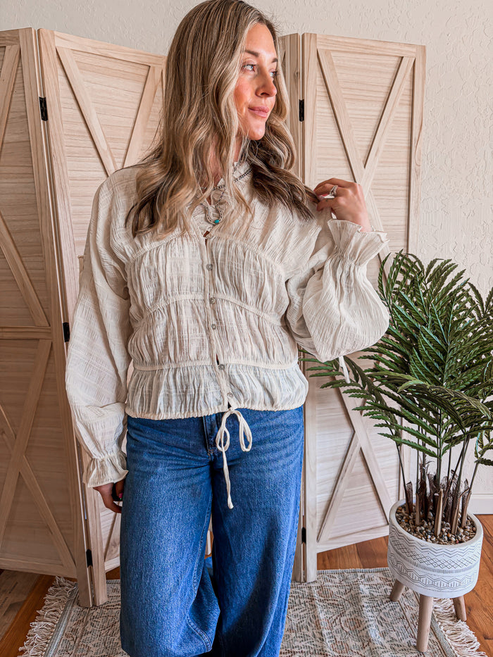 Woman wearing a cream blouse and blue jeans standing in a room with wooden shutters and a plant.