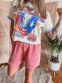 Person wearing a vintage-style t-shirt with graphic design and red checkered shorts in a casual setting.